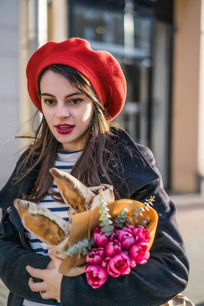 Genç güzel kız Fransız kadın esmer kırmızı bere ve siyah bir ceket Avrupa şehri Street'te bir buket çiçek ve Fransız baget ile gider. Mutlu bir gülümseme ve büyüleyici bir bak, Paris Fransız tarzı