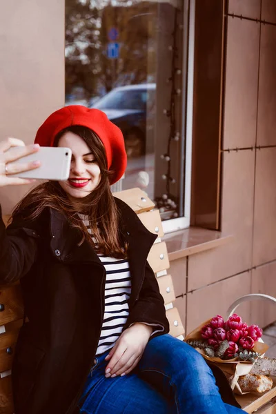beautiful brunette woman in red beret sits on the street on the bench, makes selfie, uses a smartphone