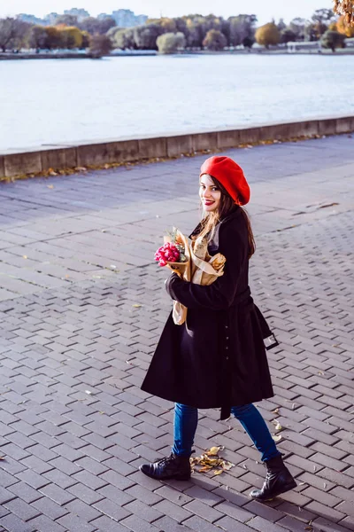 beautiful French woman brunette in a red beret with a bouquet of flowers and French baguettes under her arm walks through the park in Paris in a European city, in Paris