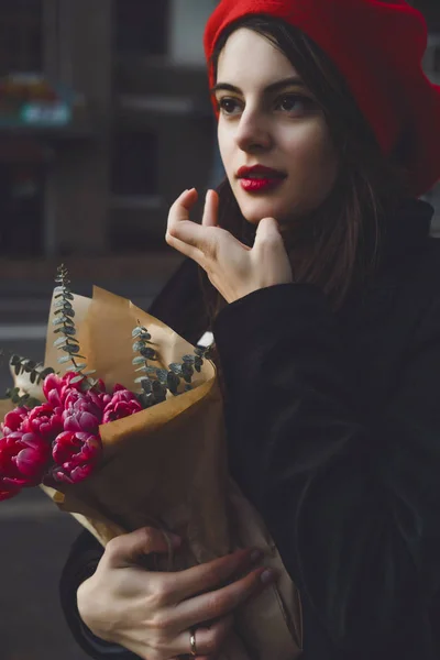 赤いベレー帽と黒いコートで美しい少女 Frenchwoman ブルネットは フランスのバゲットと花の花束とヨーロッパの都市の通りを行きます 幸せな笑顔と魅力的な外観 パリのフランス スタイル — ストック写真