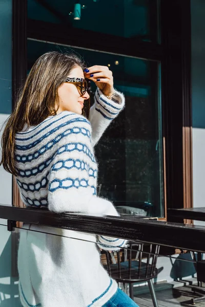 美丽的黑发女孩戴着太阳镜和舒适的针织套头衫摆在城市咖啡馆的露台上 — 图库照片