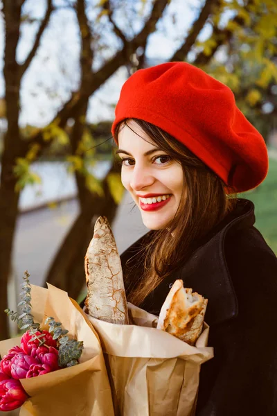 beautiful French woman brunette in a red beret with a bouquet of flowers and French baguettes under her arm walks through the park in Paris in a European city, in Paris