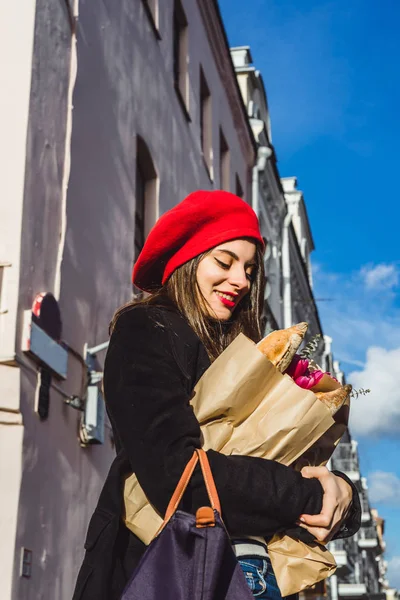 赤いベレー帽と黒いコートで美しい少女 Frenchwoman ブルネットは フランスのバゲットと花の花束とヨーロッパの都市の通りを行きます 幸せな笑顔と魅力的な外観 パリのフランス スタイル — ストック写真