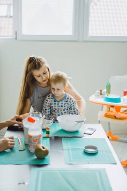 Çocuk pijamalı, annesi mutfakta elmalı turta pişiriyor ve oyuncaklarla oynuyor.