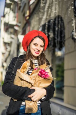 Genç güzel kız Fransız kadın esmer kırmızı bere ve siyah bir ceket Avrupa şehri Street'te bir buket çiçek ve Fransız baget ile gider. Mutlu bir gülümseme ve büyüleyici bir bak, Paris Fransız tarzı