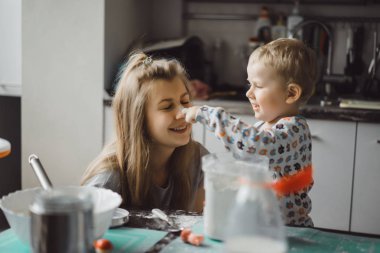 Çocuk pijamalı, annesi mutfakta elmalı turta pişiriyor ve oyuncaklarla oynuyor.