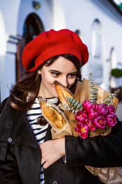 Genç güzel kız Fransız kadın esmer kırmızı bere ve siyah bir ceket Avrupa şehri Street'te bir buket çiçek ve Fransız baget ile gider. Mutlu bir gülümseme ve büyüleyici bir bak, Paris Fransız tarzı