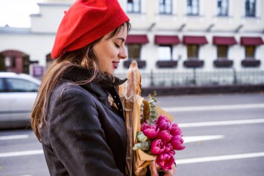 Genç güzel kız Fransız kadın esmer kırmızı bere ve siyah bir ceket Avrupa şehri Street'te bir buket çiçek ve Fransız baget ile gider. Mutlu bir gülümseme ve büyüleyici bir bak, Paris Fransız tarzı