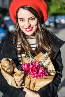 Genç güzel kız Fransız kadın esmer kırmızı bere ve siyah bir ceket Avrupa şehri Street'te bir buket çiçek ve Fransız baget ile gider. Mutlu bir gülümseme ve büyüleyici bir bak, Paris Fransız tarzı