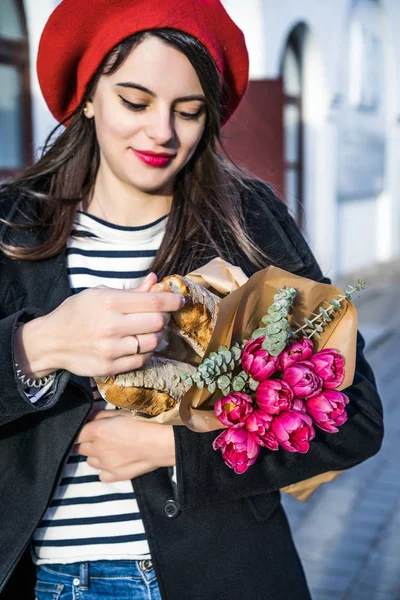 赤いベレー帽と黒いコートで美しい少女 Frenchwoman ブルネットは フランスのバゲットと花の花束とヨーロッパの都市の通りを行きます 幸せな笑顔と魅力的な外観 パリのフランス スタイル — ストック写真