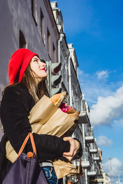 年轻美丽的女孩法国女人黑发在一个红色贝雷帽和黑色外套沿着欧洲城市的街道与花束和法国面包 一个快乐的微笑和迷人的外观 巴黎的法国风格 — 图库照片
