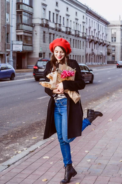 赤いベレー帽と黒いコートで美しい少女 Frenchwoman ブルネットは フランスのバゲットと花の花束とヨーロッパの都市の通りを行きます 幸せな笑顔と魅力的な外観 パリのフランス スタイル — ストック写真