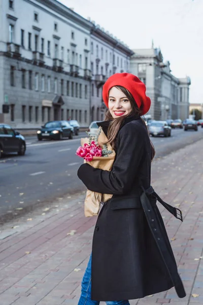 赤いベレー帽と黒いコートで美しい少女 Frenchwoman ブルネットは フランスのバゲットと花の花束とヨーロッパの都市の通りを行きます 幸せな笑顔と魅力的な外観 パリのフランス スタイル — ストック写真