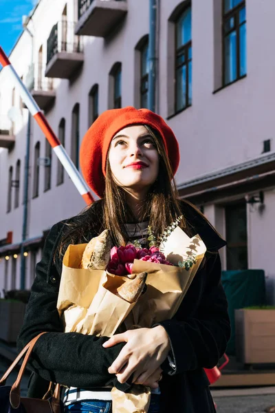 赤いベレー帽と黒いコートで美しい少女 Frenchwoman ブルネットは フランスのバゲットと花の花束とヨーロッパの都市の通りを行きます 幸せな笑顔と魅力的な外観 パリのフランス スタイル — ストック写真