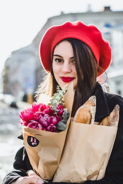 赤いベレー帽と黒いコートで美しい少女 Frenchwoman ブルネットは フランスのバゲットと花の花束とヨーロッパの都市の通りを行きます 幸せな笑顔と魅力的な外観 パリのフランス スタイル — ストック写真