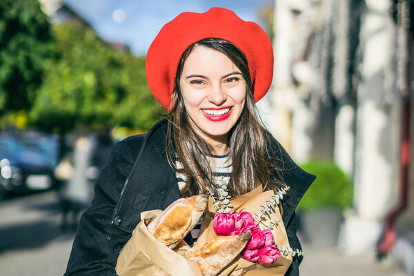 Young beautiful girl Frenchwoman brunette in a red beret and a black coat goes along the street of the European city with a bouquet of flowers and French baguettes. A happy smile and a charming look, the French style of Paris