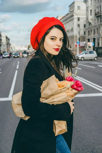 赤いベレー帽と黒いコートで美しい少女 Frenchwoman ブルネットは フランスのバゲットと花の花束とヨーロッパの都市の通りを行きます 幸せな笑顔と魅力的な外観 パリのフランス スタイル — ストック写真