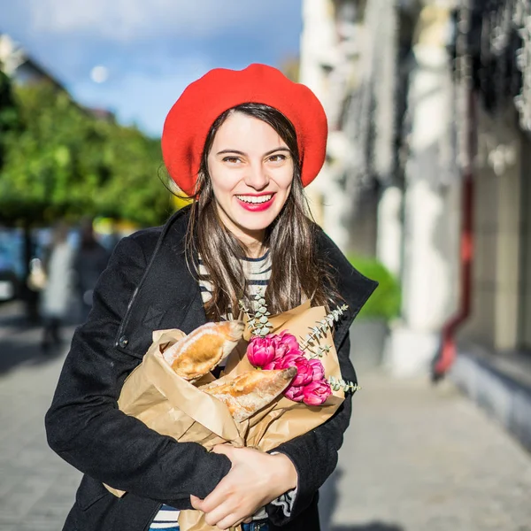 赤いベレー帽と黒いコートで美しい少女 Frenchwoman ブルネットは フランスのバゲットと花の花束とヨーロッパの都市の通りを行きます 幸せな笑顔と魅力的な外観 パリのフランス スタイル — ストック写真