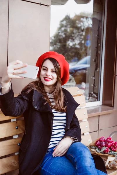 beautiful brunette woman in red beret sits on the street on the bench, makes selfie, uses a smartphone