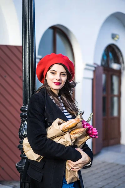 赤いベレー帽と黒いコートで美しい少女 Frenchwoman ブルネットは フランスのバゲットと花の花束とヨーロッパの都市の通りを行きます 幸せな笑顔と魅力的な外観 パリのフランス スタイル — ストック写真