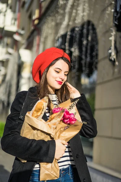 年轻美丽的女孩法国女人黑发在一个红色贝雷帽和黑色外套沿着欧洲城市的街道与花束和法国面包 一个快乐的微笑和迷人的外观 巴黎的法国风格 — 图库照片