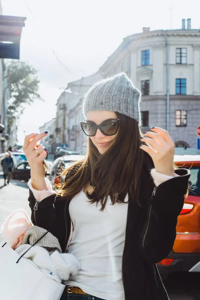 一个美丽的黑发女孩戴着太阳镜 一顶针织帽 一个迷人可爱的微笑走在欧洲城市的街道上 一个大的白色纸板包 头发发展 智能手机在她的手 — 图库照片