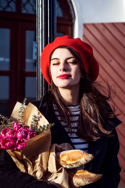 赤いベレー帽と黒いコートで美しい少女 Frenchwoman ブルネットは フランスのバゲットと花の花束とヨーロッパの都市の通りを行きます 幸せな笑顔と魅力的な外観 パリのフランス スタイル — ストック写真