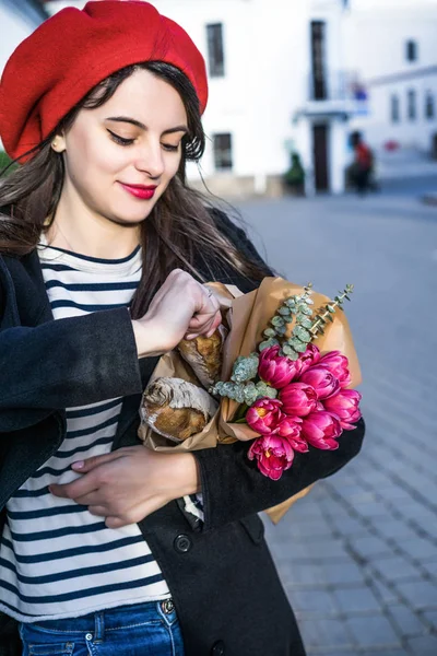 赤いベレー帽と黒いコートで美しい少女 Frenchwoman ブルネットは フランスのバゲットと花の花束とヨーロッパの都市の通りを行きます 幸せな笑顔と魅力的な外観 パリのフランス スタイル — ストック写真