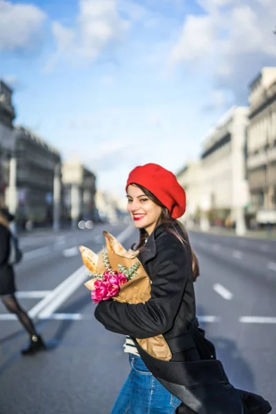 赤いベレー帽と黒いコートで美しい少女 Frenchwoman ブルネットは フランスのバゲットと花の花束とヨーロッパの都市の通りを行きます 幸せな笑顔と魅力的な外観 パリのフランス スタイル — ストック写真