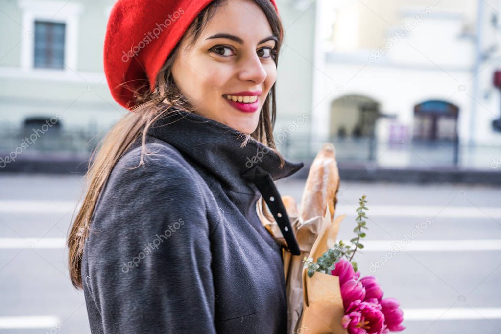 Young beautiful girl Frenchwoman brunette in a red beret and a black coat goes along the street of the European city with a bouquet of flowers and French baguettes. A happy smile and a charming look, the French style of Paris