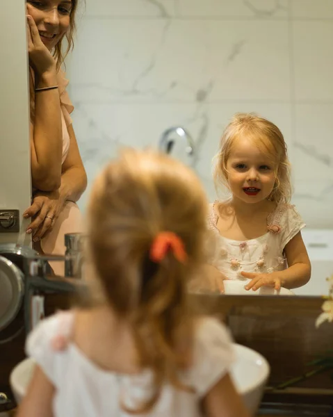 Mom and daughters do makeup in the bathroom, apply lipstick in front of the mirror. Mom and daughters have fun, spend time together.