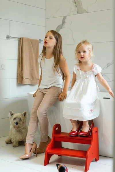 Mom and daughters do makeup in the bathroom, apply lipstick in front of the mirror. Daughters in her mother\'s heels. Mom and daughters have fun, spend time together.