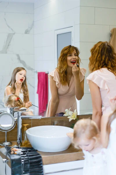 Mom and daughters do makeup in the bathroom, apply lipstick in front of the mirror. Mom and daughters have fun, spend time together.
