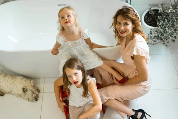 Mom and daughters do makeup in the bathroom, apply lipstick in front of the mirror. Daughters in her mother\'s heels. Mom and daughters have fun, spend time together.