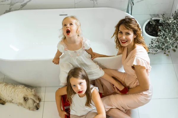 Mom and daughters do makeup in the bathroom, apply lipstick in front of the mirror. Daughters in her mother\'s heels. Mom and daughters have fun, spend time together.