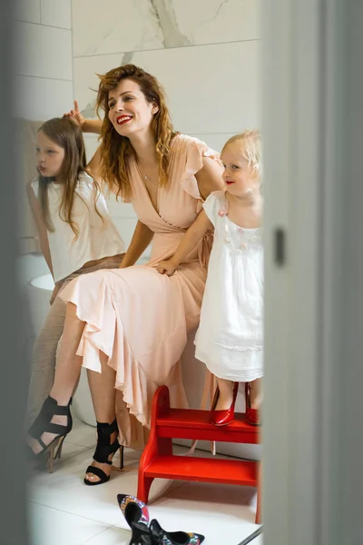 Mom and daughters do makeup in the bathroom, apply lipstick in front of the mirror. Daughters in her mother\'s heels. Mom and daughters have fun, spend time together.