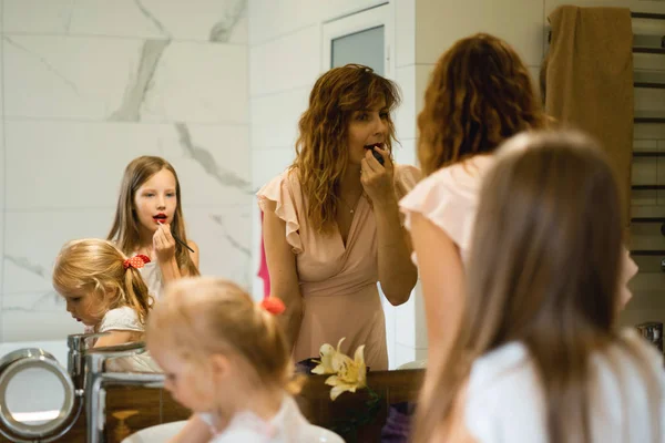 Mom and daughters do makeup in the bathroom, apply lipstick in front of the mirror. Mom and daughters have fun, spend time together.