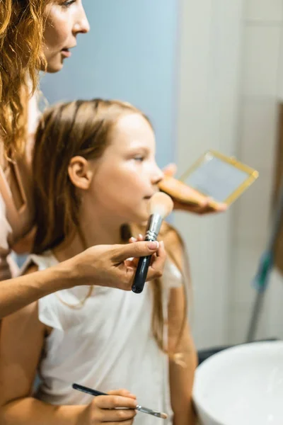 Mom and daughters do makeup in the bathroom, apply lipstick in front of the mirror. Mom and daughters have fun, spend time together.