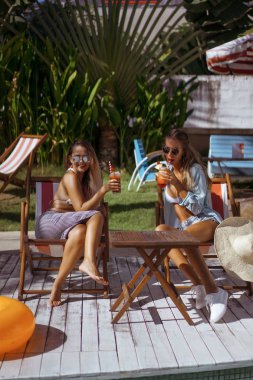 iki genç kadın arkadaş bir kafede Kahvaltı, haberlerde tartışırken, gülüyor. kafede kadın havuz kenarında şezlong üzerinde kokteyl içmek. iki çekici genç kadın havuz kenarında şezlong dinlenirken kokteyl içme bikini