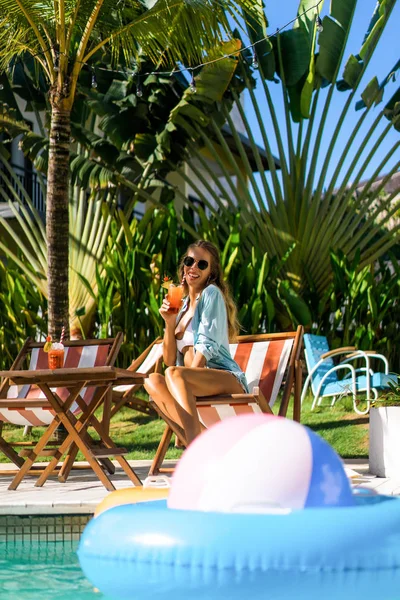 two young women friends at a cafe having breakfast, discussing the news, laughing. females in the cafe drink cocktails on the loungers by the pool. two attractive young women in bikini drinking cocktails while relaxing in deck chair near the pool