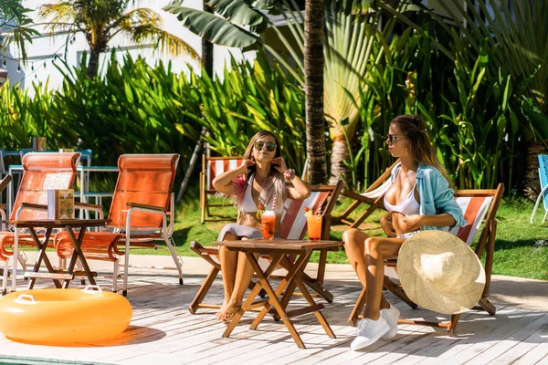 two young women friends at a cafe having breakfast, discussing the news, laughing. females in the cafe drink cocktails on the loungers by the pool. two attractive young women in bikini drinking cocktails while relaxing in deck chair near the pool