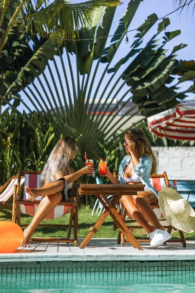 two young women friends at a cafe having breakfast, discussing the news, laughing. females in the cafe drink cocktails on the loungers by the pool. two attractive young women in bikini drinking cocktails while relaxing in deck chair near the pool