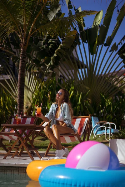 two young women friends at a cafe having breakfast, discussing the news, laughing. females in the cafe drink cocktails on the loungers by the pool. two attractive young women in bikini drinking cocktails while relaxing in deck chair near the pool