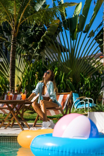 two young women friends at a cafe having breakfast, discussing the news, laughing. females in the cafe drink cocktails on the loungers by the pool. two attractive young women in bikini drinking cocktails while relaxing in deck chair near the pool