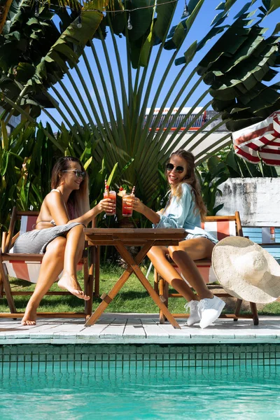 iki genç kadın arkadaş bir kafede Kahvaltı, haberlerde tartışırken, gülüyor. kafede kadın havuz kenarında şezlong üzerinde kokteyl içmek. iki çekici genç kadın havuz kenarında şezlong dinlenirken kokteyl içme bikini