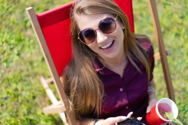 Akıllı telefonu olan ve güverte sandalyesinde doğada kokteyl içen bir kız. Yaz tatili.