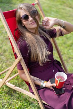 Akıllı telefonu olan ve güverte sandalyesinde doğada kokteyl içen bir kız. Yaz tatili.