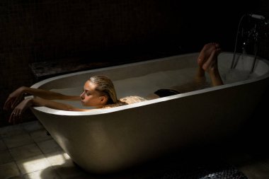 Güzel gülümseyen beyaz kadın banyoda ıslak saçlarıyla poz veriyor, banyoda dinleniyor. Genç bir kadın küvette yatıyor..