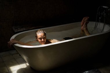 Güzel gülümseyen beyaz kadın banyoda ıslak saçlarıyla poz veriyor, banyoda dinleniyor. Genç bir kadın küvette yatıyor..