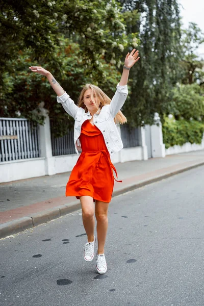 赤いドレスと白いデニムのジャケットを着た美しい若い女性が街を歩き回って幸せな笑顔 — ストック写真